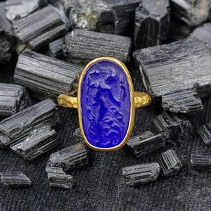 Può includere: Un anello in oro con una pietra preziosa di lapislazzuli blu intenso di forma ovale e un intricato design intagliato. La fascia in metallo presenta una finitura martellata e testurizzata che contrasta con la superficie liscia della pietra blu.