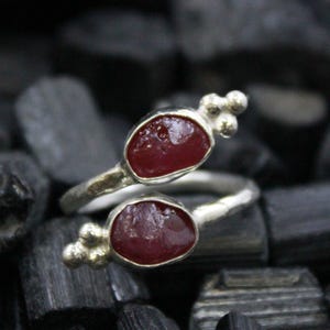 May include: A silver ring with two oval, deep red gemstones. The ring has a unique, open design with small, round silver accents near the gemstones. The ring is set against a dark, textured background.