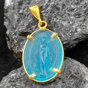 Puede incluir: Un colgante de oro con un camafeo de vidrio azul de la Virgen Mar&iacute;a. El camafeo tiene forma ovalada y tiene un marco de oro.
