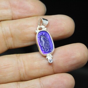 May include: Silver pendant with a purple oval-shaped stone featuring a carved figure. The pendant has a small loop at the top and a small, round, clear stone at the bottom. The figure is a woman with flowing hair and drapery.