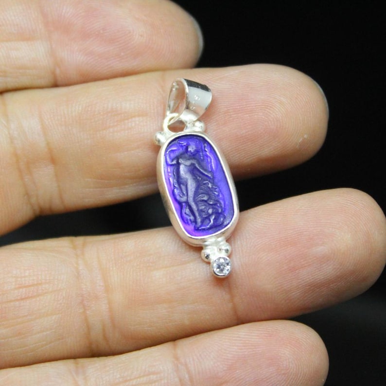 May include: Silver pendant with a purple oval-shaped stone featuring a carved figure. The pendant has a small loop at the top and a small, round, clear stone at the bottom. The figure is a woman with flowing hair and drapery.