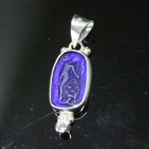 May include: A silver pendant with a rectangular, deep blue center featuring a carved figure. The pendant has a bail at the top and a small, clear gemstone at the bottom. The design is likely inspired by classical art.