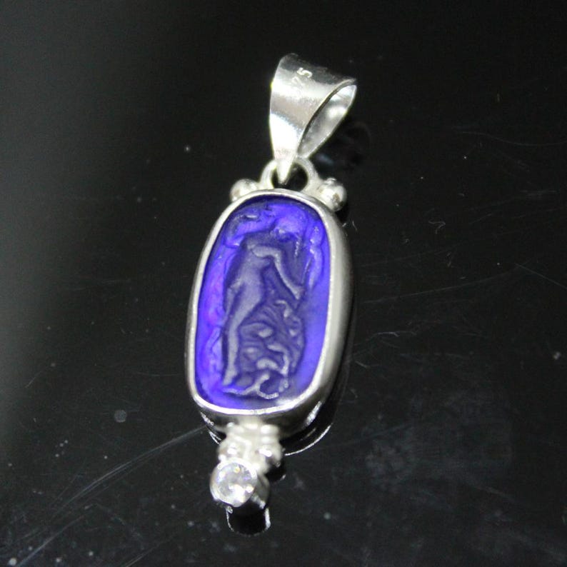 May include: A silver pendant with a rectangular, deep blue center featuring a carved figure. The pendant has a bail at the top and a small, clear gemstone at the bottom. The design is likely inspired by classical art.
