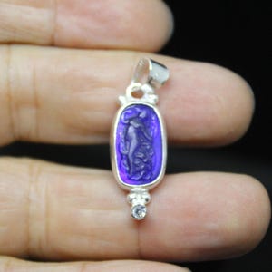 May include: A silver pendant with a rectangular, purple-colored center featuring a carved figure. The pendant has a small, clear gemstone at the bottom and a silver loop at the top for a chain. The figure is a classical female form.