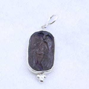 May include: A silver pendant with a dark purple, rectangular stone. The stone features a carved image of a winged figure. The pendant has a loop at the top and a decorative cluster of three small spheres at the bottom.