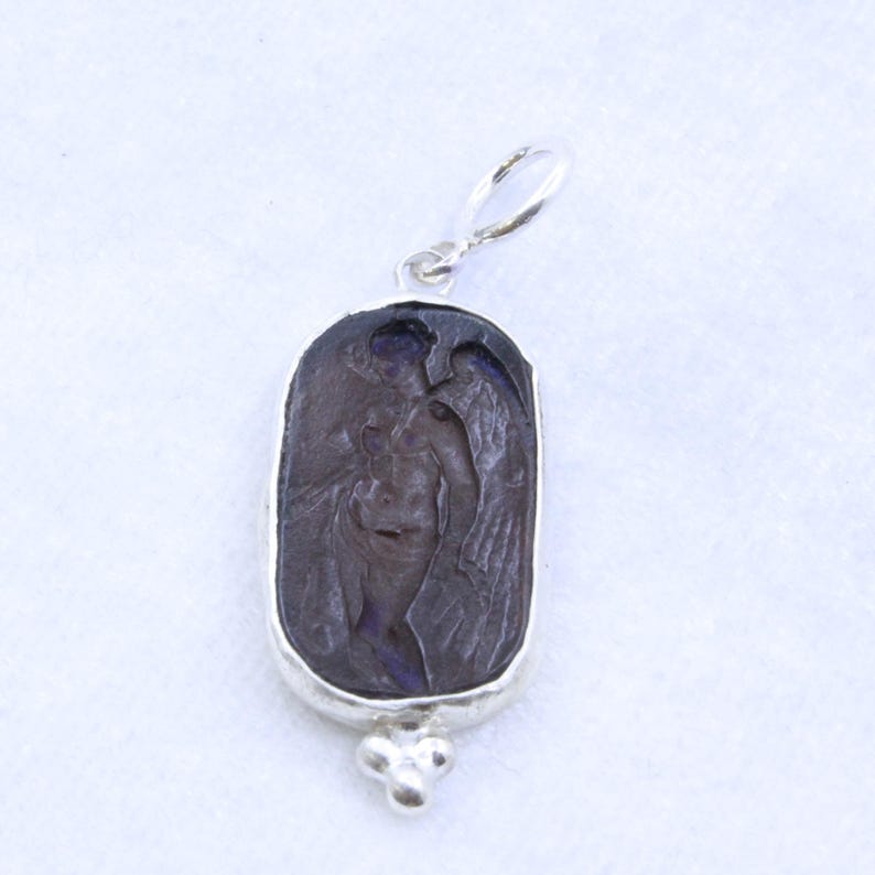 May include: A silver pendant with a dark purple, rectangular stone. The stone features a carved image of a winged figure. The pendant has a loop at the top and a decorative cluster of three small spheres at the bottom.