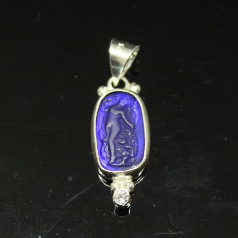 May include: A silver pendant with a blue oval-shaped center featuring a carved figure. The pendant has a small, clear gemstone at the bottom and a silver loop at the top for a chain. The background is black.