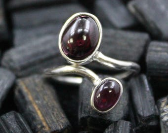 May include: A silver ring featuring two oval, deep red gemstones. The gemstones are set in a silver band that curves around the finger. The ring is displayed on a bed of black stones.