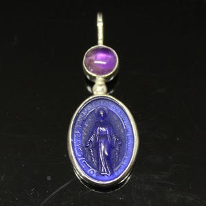 May include: A silver pendant featuring a blue oval with a religious figure and a purple gemstone. The oval pendant has a silver border and a detailed depiction of a religious figure. The gemstone is round and purple.