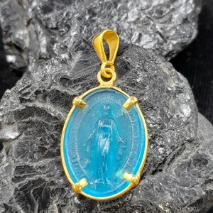 Puede incluir: Pendiente dorado con un camafeo ovalado de vidrio azul de la Virgen Mar&iacute;a. El camafeo est&aacute; rodeado de un marco dorado con detalles decorativos.