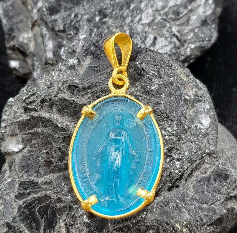 Puede incluir: Pendiente dorado con un camafeo ovalado de vidrio azul de la Virgen Mar&iacute;a. El camafeo est&aacute; rodeado de un marco dorado con detalles decorativos.