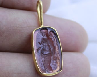 May include: A gold pendant with a rectangular, translucent, purple gemstone featuring a carved image of an angel with wings.