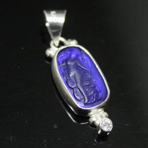 May include: A silver pendant with a deep blue, oval-shaped center. The blue center features a carved image of a figure. A small, clear gemstone is set at the bottom. The pendant has a bail for a chain.