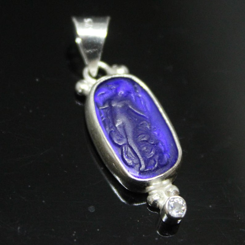 May include: A silver pendant with a deep blue, oval-shaped center. The blue center features a carved image of a figure. A small, clear gemstone is set at the bottom. The pendant has a bail for a chain.