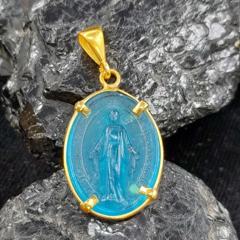Puede incluir: Pendiente de oro con un &oacute;valo de vidrio azul que representa a la Virgen Mar&iacute;a. El pendiente tiene un aro de oro y cuatro acentos dorados alrededor del &oacute;valo.