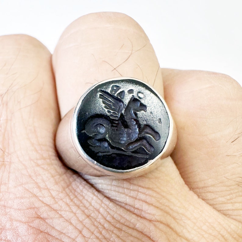 May include: A silver ring with a black onyx stone featuring a carved image of a winged horse.