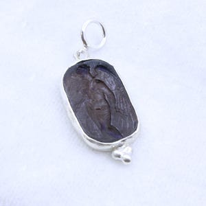 May include: A silver pendant with a dark, rectangular stone featuring a carved figure with wings. The stone is set in a silver bezel and has a small loop at the top for a chain. A cluster of three small silver beads is at the bottom.
