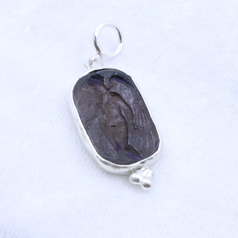 May include: A silver pendant with a dark, rectangular stone featuring a carved figure with wings. The stone is set in a silver bezel and has a small loop at the top for a chain. A cluster of three small silver beads is at the bottom.