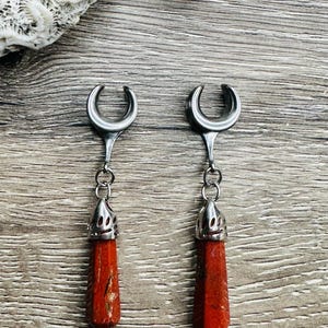 May include: A pair of silver crescent moon ear gauges with red stone dangles. The stones are faceted and have a reddish-brown color.