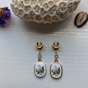 Op de afbeelding: Een paar goudkleurige oorplugs met halvemaanvormige bovenkanten en bungelende ovale hangers. Elke hanger heeft een witte achtergrond met een gedetailleerde illustratie van een sneeuwuil die op groene dennentakken zit, omlijst door dun metaal.