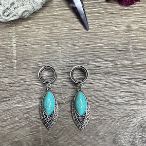 May include: A pair of silver-toned ear gauges with turquoise teardrop-shaped pendants. The pendants have a detailed, ornate silver frame. The gauges are displayed on a wooden surface.
