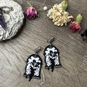 Pode incluir: Um par de brincos preto e branco com um design de arco gótico com uma figura dentro. Os brincos são presos a postes prateados.