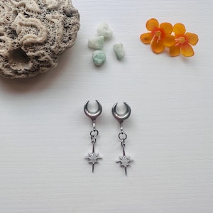 May include: A pair of silver-toned earrings with a crescent moon design at the top and a dangling star pendant. The star pendant has a small, round, white stone in the center. The earrings are displayed on a white surface, with coral, small green stones, and orange flowers.