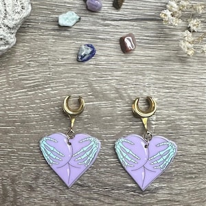 Puede incluir: Un par de pendientes de corazón dorados con un diseño de mano esquelética y un degradado morado y azul. Los pendientes están diseñados para ser usados como piercing corporal.