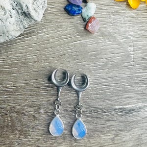 May include: Silver-coloured earrings with a crescent moon top and a chain leading to a teardrop-shaped, opalescent stone. The earrings are displayed on a wooden surface, with colourful gemstones and a yellow flower in the background.