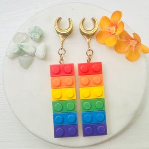 May include: A pair of gold-colored tunnel earrings with rainbow-colored brick pendants. The pendants are made of red, orange, yellow, green, blue, and purple blocks. The earrings are displayed on a white marble surface with small green and white stones and orange flowers.