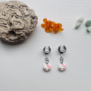 May include: A pair of silver-toned earrings with a crescent-shaped top and a dangling Hello Kitty charm. The charm is white with a pink bow and whiskers. The earrings are displayed on a white surface with coral and orange flowers.