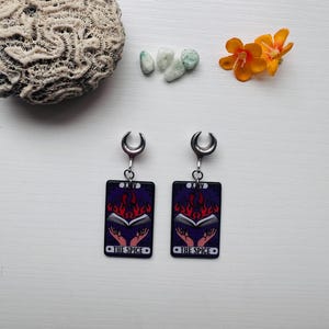 May include: A pair of silver-toned earrings with a black rectangular charm. The charm features a tarot card design with a book, flames, and hands, with the text "THE SPICE". The earrings are displayed with a coral, small stones, and orange flowers.