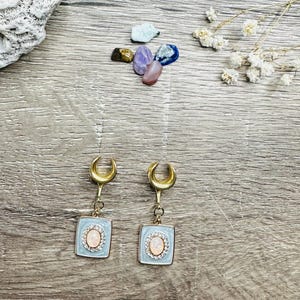 Puede incluir: Un par de expansores de oreja en forma de media luna dorada con un encanto cuadrado blanco y azul claro con una ópalo rosa.