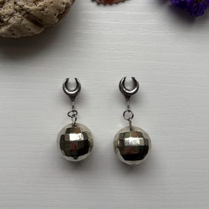 May include: A pair of silver-toned earrings featuring a crescent-shaped top and a faceted, spherical charm. The earrings are suspended from a small chain. The background includes a seashell, a piece of coral, and purple flowers.
