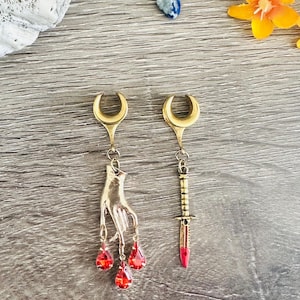 Puede incluir: Un par de expansores de oreja de media luna dorados con colgantes. Un colgante es una mano dorada con tres gotas de cristal rojo. El otro colgante es una daga dorada con una punta roja.