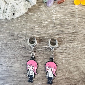 May include: A pair of silver crescent moon earrings with a black and pink anime character charm. The character has pink hair, a white shirt, and black pants.