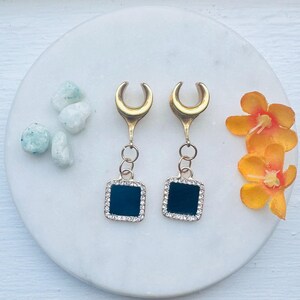 May include: A pair of gold-colored ear gauges with a dark teal square pendant outlined with small, clear crystals. The gauges are displayed on a white marble surface, with small decorative stones and orange flowers.