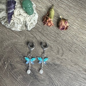 May include: A pair of silver crescent moon earrings with blue and purple dragonfly charms and white pearl accents.