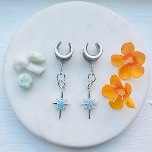 Puede incluir: Un par de expansores de oreja plateados con un diseño de estrella y una piedra central azul claro. Los expansores se exhiben sobre una superficie de mármol blanco, con flores naranjas y pequeñas piedras verdes.