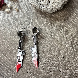 May include: Two silver ear gauges with a bloody knife design. The knife has a black handle and a white blade with a red blood stain.