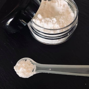May include: A white powder in a clear jar with a black lid. A small plastic spoon with a scoop of the powder is next to the jar.