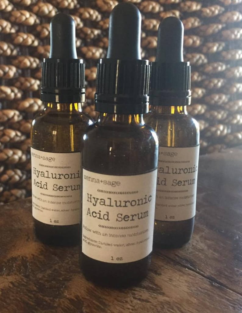Może przedstawiać: Trzy brązowe butelki szklane z czarnymi zakraplaczami i białymi etykietami. Etykiety zawierają napis "Hyaluronic Acid Serum" i "enna+sage". Każda butelka zawiera 1 uncję serum.