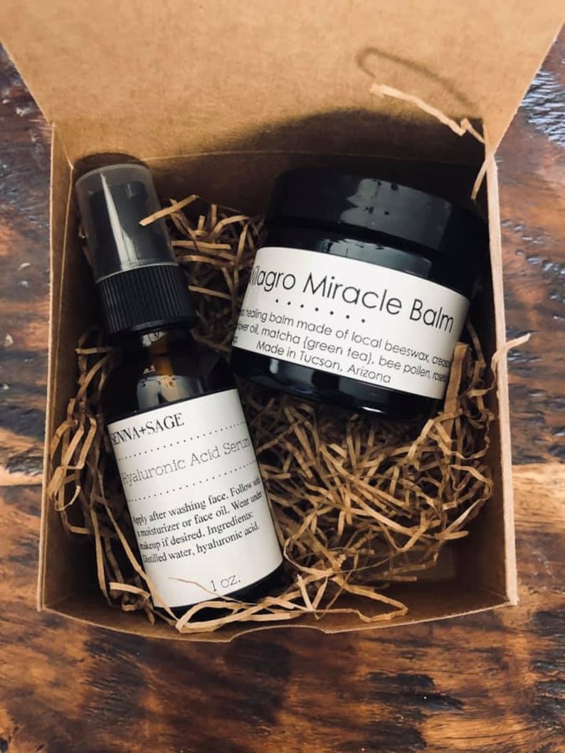 May include: A small cardboard box with brown shredded paper inside. The box contains a small bottle of hyaluronic acid serum and a jar of milagro miracle balm. The serum bottle is labeled "ENNA-SAGE Hyaluronic Acid Serum" and the balm jar is labeled "Magro Miracle Balm".