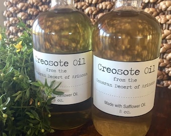May include: Two clear glass bottles with black lids filled with a light yellow liquid. The bottles have white labels with black text that reads "Creosote Oil from the Sonoran Desert of Arizona" and "Made with Safflower Oil. 8 oz."
