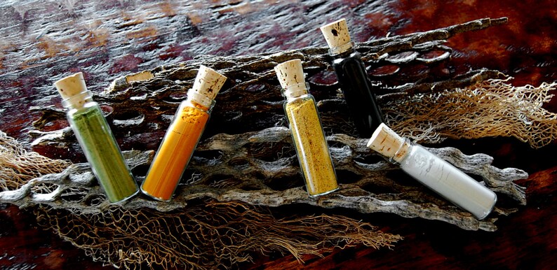 May include: Five small glass bottles with cork tops, filled with different colored powders. The bottles are arranged in a row on a wooden surface with a piece of dried seaweed behind them.
