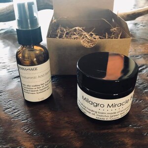 May include: A set of two skincare products in a brown cardboard box with shredded paper. The products are a small glass bottle with a black pump top and a small black jar with a white label that reads "Milagro Miracle Balm". The bottle label reads "ENNA+SAGE Hyaluronic Acid Serum".