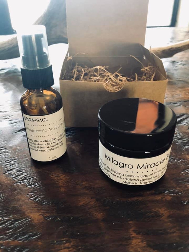 May include: A set of two skincare products in a brown cardboard box with shredded paper. The products are a small glass bottle with a black pump top and a small black jar with a white label that reads "Milagro Miracle Balm". The bottle label reads "ENNA+SAGE Hyaluronic Acid Serum".