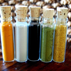May include: Five small glass bottles with cork tops, each filled with a different colored powder. The powders are yellow, white, black, green, and yellow.