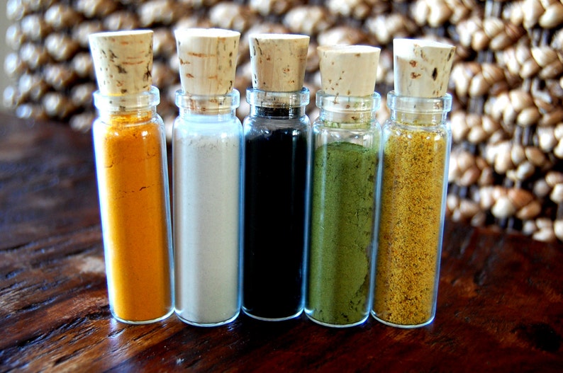 May include: Five small glass bottles with cork tops, each filled with a different colored powder. The powders are yellow, white, black, green, and yellow.