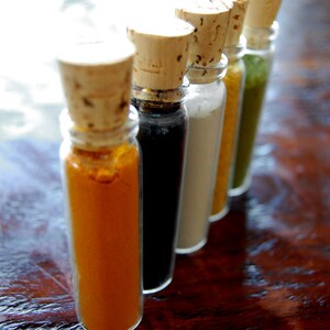 May include: Five small glass bottles with cork tops, filled with different colored powders. The bottles are lined up in a row on a wooden surface. The powders are yellow, black, white, light brown, and green.